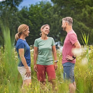 Three people in casual summer clothing standing in a wildflower meadow with tall grass and flowers, surrounded by green trees under blue sky.