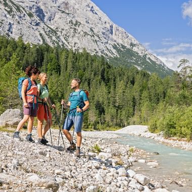Three hikers with backpacks and trekking poles standing on rocky riverbank of Leutascher Ache in Bavaria, with mountain and forest backdrop.