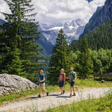 Drei Wanderer mit Rucksäcken auf einem Bergweg beim Gosausee mit schneebedeckten Gipfeln, grünen Wäldern und einem See im Hintergrund.
