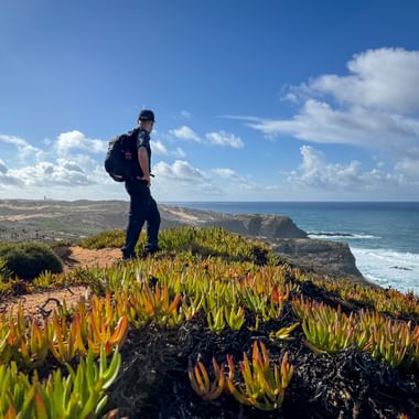 Hiker with backpack standing on cliff with colorful succulent plants overlooking the Atlantic Ocean on the Rota Vicentina trail in Portugal.