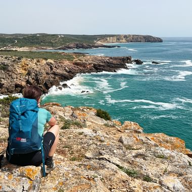Hiker with blue backpack sitting on rocky cliff overlooking turquoise Atlantic Ocean at Praia da Ingrina, Algarve coastline with dramatic cliffs.