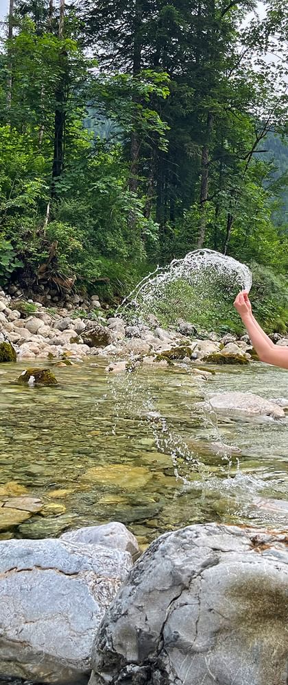 Hiker with backpack crouching on rocks in Lienbach mountain stream at Postalm, splashing water, surrounded by forest and mountains.