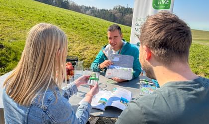Eurohike guide in turquoise jacket discussing hiking maps with two guests at outdoor table. Green meadow and forest in background with Eurohike banner.