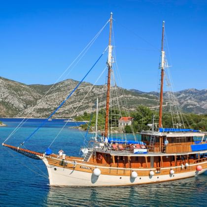 Traditional wooden sailing ship MS Sagena with two masts in turquoise waters of Kvarner Bay, Croatia, with mountains and coastline in background.