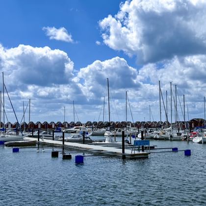 Hafen in Träslövsläge mit Segelbooten an Stegen. Rote Holzbootshäuser säumen das Ufer unter blauem Himmel mit weißen Wolken.