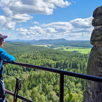 Wanderer mit blauem Rucksack am Pfaffenstein Aussichtspunkt mit Blick auf bewaldete Täler und Felder. Markante Felsformation neben dem Geländer.
