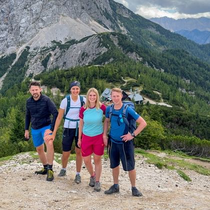 Vier Wanderer mit Rucksäcken auf dem Vršič-Pass-Gipfel mit bewaldeten Bergen und felsigen Gipfeln der Julischen Alpen im Hintergrund.