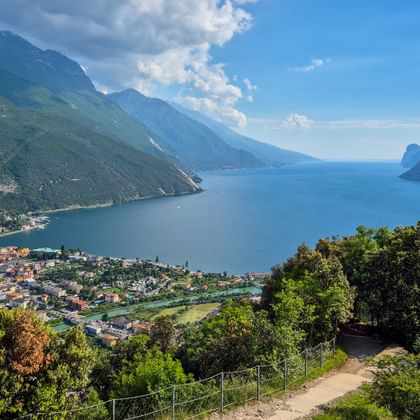 Hiking trail on Monte Brione overlooking Lake Garda, Torbole, and surrounding mountains under blue sky with white clouds.