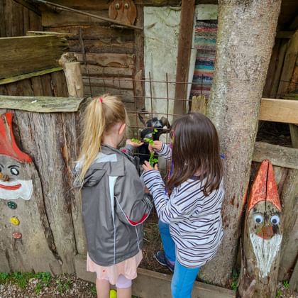 Zwei Kinder füttern Ziegen durch einen Holzzaun in einem rustikalen Stall. Bemalte Zwerge schmücken die verwitterte Holzstruktur.