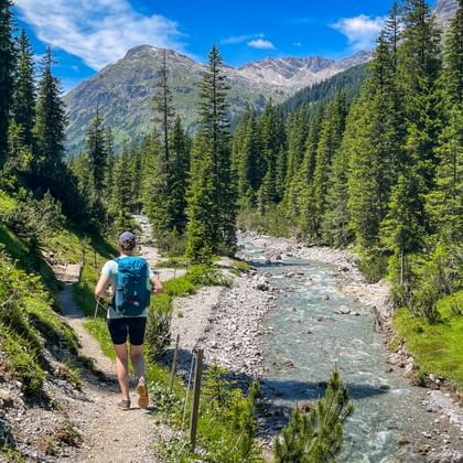 Hiker with backpack walking on trail beside mountain stream. Dense conifer forest lines both sides, with rocky peaks under blue sky.