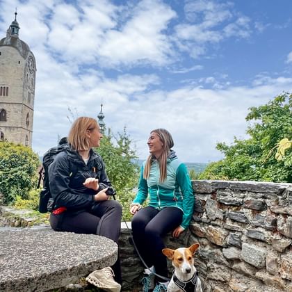 Two women hikers with a dog resting on a stone wall in Stein an der Donau, Wachau. Historic church tower and vineyard landscape visible.