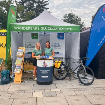 Two staff members in turquoise shirts at an Eurobike promotional tent with banners and a bicycle. Green banner reads 'Wanderbare Glücksmomente'.