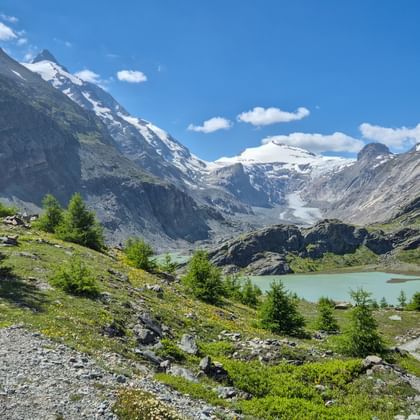 Alpe Adria Trail Wegweiser beim Margaritzensee mit Großglockner Gletscher und schneebedeckten Gipfeln unter blauem Himmel.