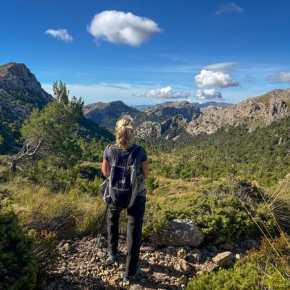 Hiker with backpack standing on rocky trail overlooking Tramuntana mountain range with green valleys and blue sky with white clouds.