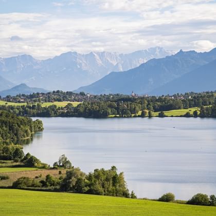 Scenic view of a lake surrounded by green meadows and forests with snow-capped Alpine mountains in the background under a cloudy sky.