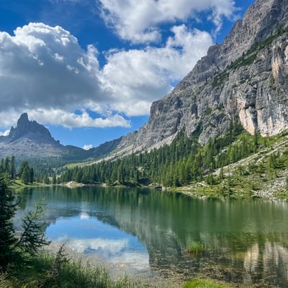 Lago Federa spiegelt dramatische Dolomitengipfel und Wolken. Tannenwälder umgeben den grünen Alpensee mit steilen Felsklippen rechts.
