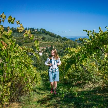 Frau wandert durch grüne Weinbergreihen in der Toskana mit zypressengesäumten Hügeln und historischen Gebäuden im Hintergrund unter blauem Himmel.