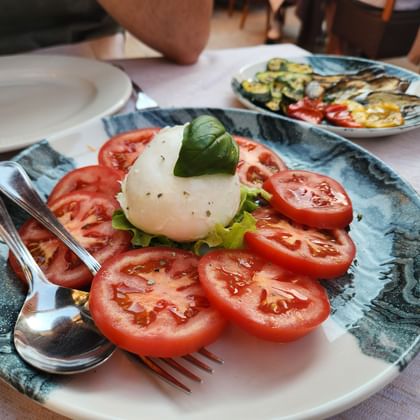 Caprese salad with sliced tomatoes, mozzarella, and basil on a blue plate. Grilled vegetables visible on a second plate in the background.