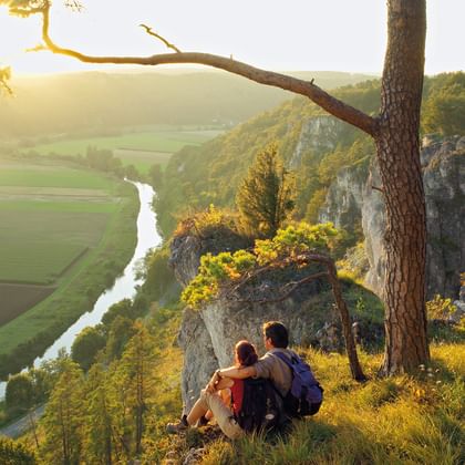 Two hikers sitting on a rocky cliff in Altmühltal, overlooking a winding river through green valley at sunset with pine tree in foreground.