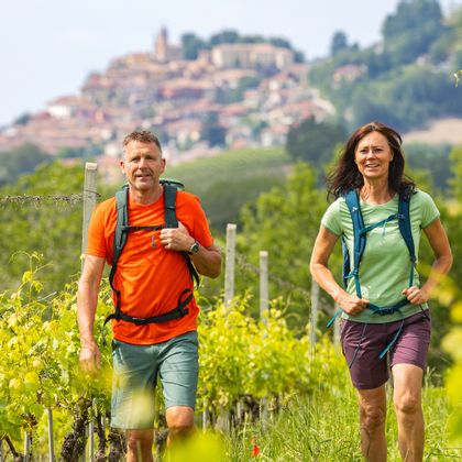 Zwei Wanderer zwischen Weinreben mit dem Ort Montforte im Hintergrund