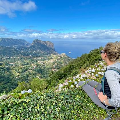 Female hiker with blue backpack sitting on hillside overlooking Madeira's north coast with dramatic cliffs, ocean, and white flowers in foreground.