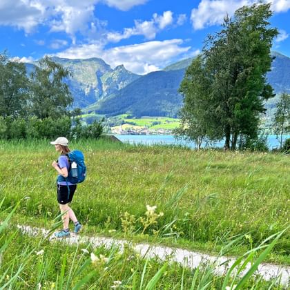 Wanderer mit blauem Rucksack auf Wiesenweg im Salzkammergut. Türkisfarbener See und Berge im Hintergrund unter blauem Himmel.