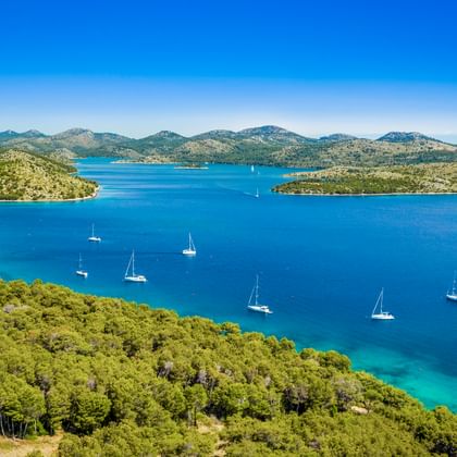 Aerial view of Dugi Otok bay in North Dalmatia with turquoise water, white sailboats, green forested coastline, and mountains under blue sky.