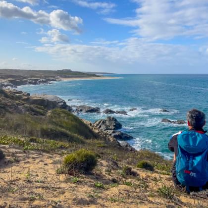 Wanderer mit blauem Rucksack sitzt auf Küstenklippe mit Blick auf türkisfarbenen Atlantik bei Porto Covo, Praia do Sissal, Rota Vicentina, Portugal.