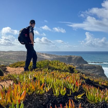 Hiker with backpack standing on cliff with succulent plants overlooking Atlantic Ocean on Rota Vicentina trail in Portugal.