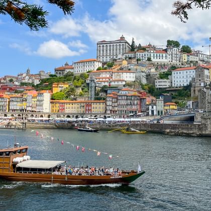 Traditionelles Holzboot mit Passagieren auf dem Douro in Porto, mit bunten Häusern am Hang und der Dom Luís I Brücke.