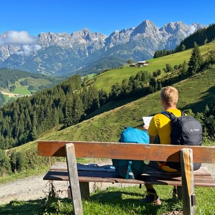 Two hikers with backpacks sit on a wooden bench overlooking green alpine meadows and the rocky Kitzbüheler Alps under a blue sky.