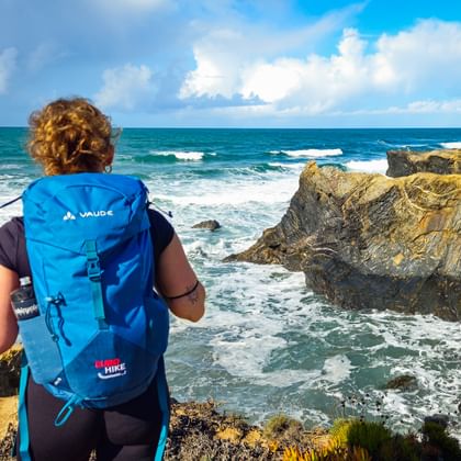 Wanderin mit blauem Rucksack betrachtet felsige Küste an der Praia das Furnas an der Rota Vicentina. Wellen brechen an goldenen Felsen unter blauem Himmel.