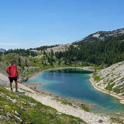 Hiker in red jacket overlooking turquoise alpine lake in Julian Alps valley, surrounded by rocky slopes, green vegetation and mountain peaks.