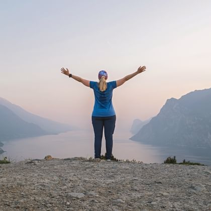 Woman with raised arms standing on rocky summit at Monte Brione overlooking Lake Garda at sunset, mountains visible on both sides.