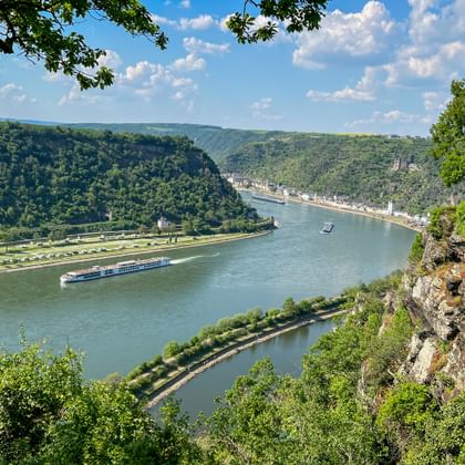 Panoramic view of Rhine River winding through green valley with cruise ship, forested hills, and town along riverbank, seen from Loreley Rock.