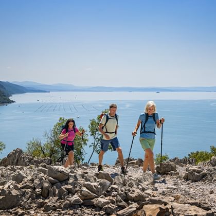 Three hikers with backpacks and trekking poles on rocky trail overlooking the blue Adriatic Sea near Trieste, Italy.