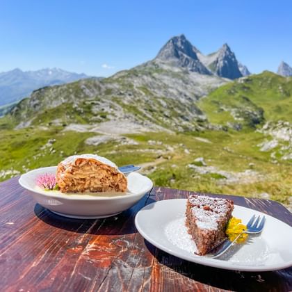 Two dessert slices on white plates at a mountain hut table, with green alpine meadows and rocky peaks of the Allgäu-Lechtal Alps behind.