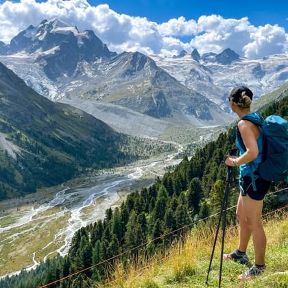 Hiker enjoying the view of the valley to Pontresina, with a meandering stream and the snow-covered peaks in the background
