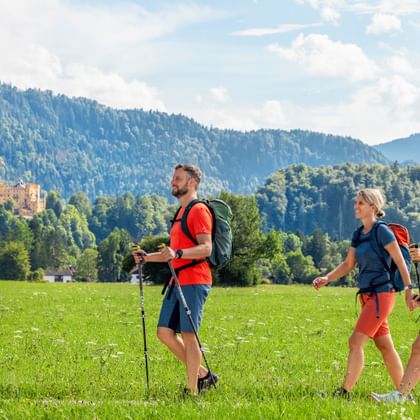 Drei Wanderer mit Rucksäcken gehen über eine grüne Wiese bei Hohenschwangau. Bewaldete Berge und Schloss Hohenschwangau im Hintergrund sichtbar.