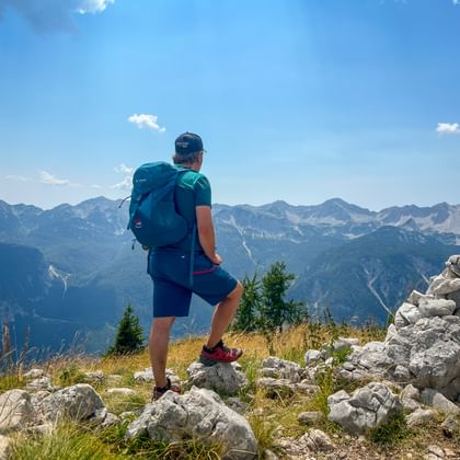 Hiker with backpack standing on rocky summit in the Julian Alps, overlooking mountain ranges under blue sky with stone cairn nearby.
