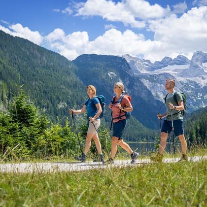 Drei Wanderer mit Rucksäcken auf einem Weg am Gosausee mit dem schneebedeckten Dachstein-Gebirge und bewaldeten Hängen im Hintergrund.