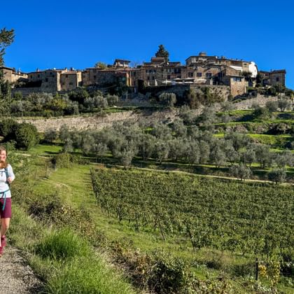 Wanderin mit Rucksack auf Schotterweg durch grünen Hang mit Olivenbäumen. Historisches Steindorf auf Hügel unter blauem Himmel sichtbar.