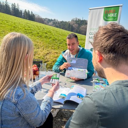 Eurohike-Guide in türkisfarbener Jacke bespricht Wanderkarten mit zwei Gästen an Tisch im Freien. Grüne Wiese und Wald im Hintergrund mit Eurohike-Banner.