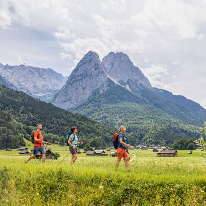 Three hikers with backpacks walking through green meadows near Garmisch with dramatic Alpine peaks in the background under cloudy sky.