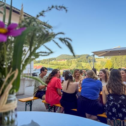 Mitarbeiter sitzen an Biergarnituren bei einem Sommerfest im Freien. Rosa Blumen im Vordergrund, Hügel und blauer Himmel im Hintergrund.