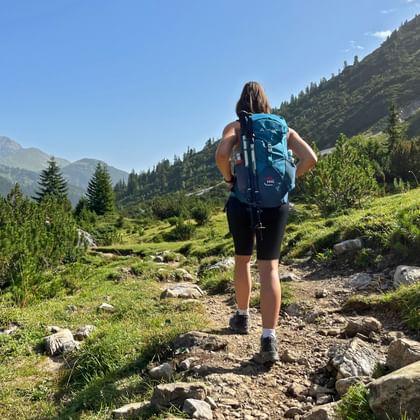 Wanderer mit blauem Rucksack auf felsigem Bergweg durch grünes Alpental mit bewaldeten Hängen und Berggipfeln unter blauem Himmel.