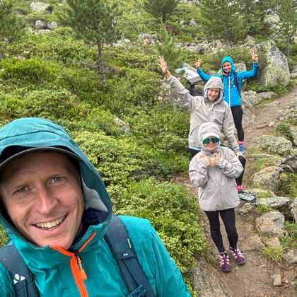 Smiling man taking selfie with family on rocky mountain trail in rain. Three people behind wear rain jackets, surrounded by green vegetation.