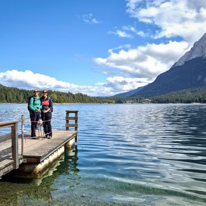 Zwei Wanderer stehen auf einem Holzsteg am Eibsee mit klarem türkisfarbenem Wasser, bewaldeten Ufern und der Zugspitze im Hintergrund.