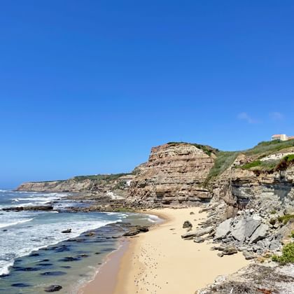 Sandy beach at São Pedro da Cadeira bay with layered rock cliffs, waves breaking on shore, and buildings visible on hilltop under blue sky.