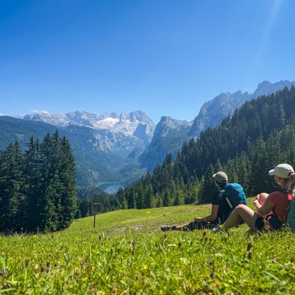 Two hikers with backpacks sitting on a green meadow in Gosau, viewing the Dachstein glacier and mountain peaks under a clear blue sky.